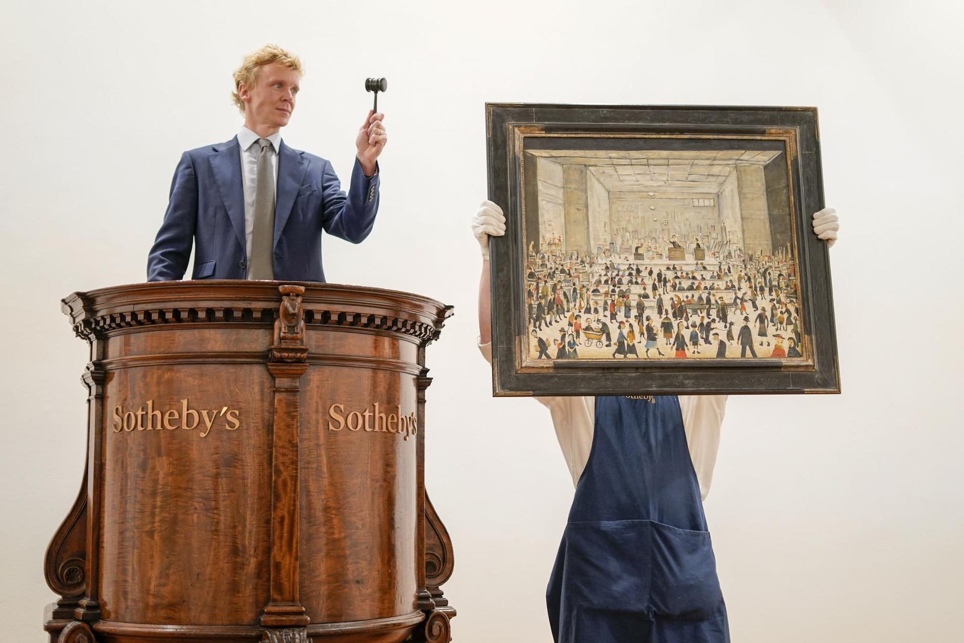 Một nhân viên giữ bức tranh 'The Auction', bức tranh duy nhất được biết đến trong phòng đấu giá của họa sĩ người Anh LS Lowry, tại hội trường đấu giá Sotheby's, ở thủ đô London, vương quốc Anh. Ảnh: AP/Alberto Pezzali.